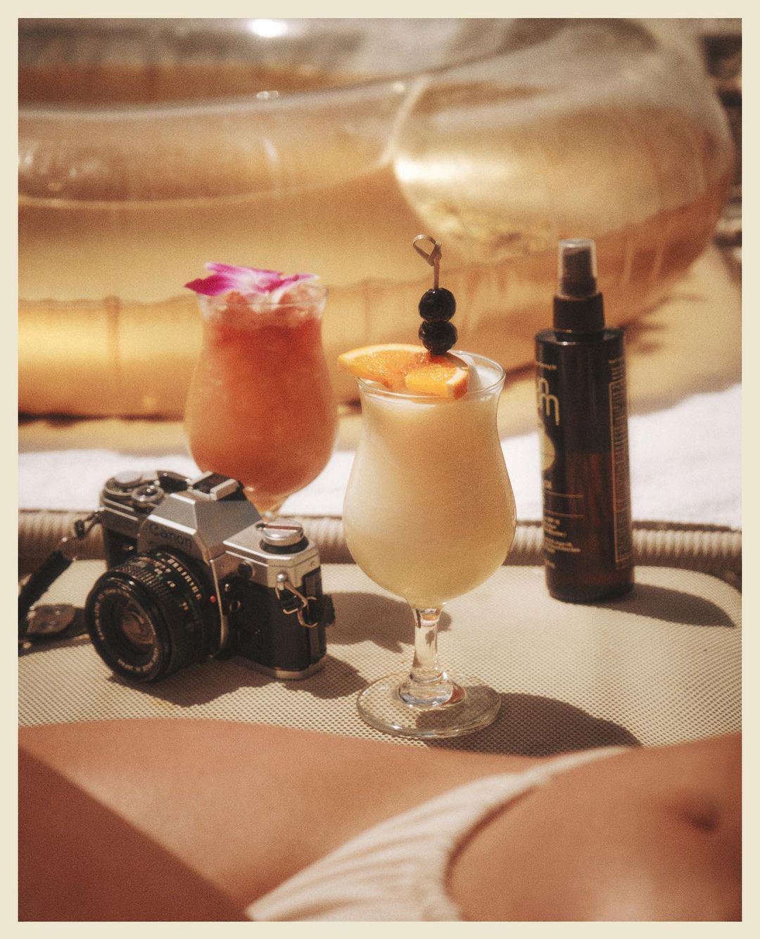 Two tropical cocktails beside a vintage camera and a bottle of sunscreen on a tan surface, with an inflatable pool in the background.