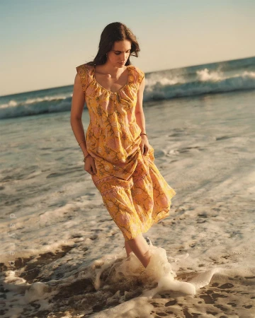 A woman in a yellow floral dress stands in the ocean surf at the beach in a medium shot on a sunny day with blue ocean background.