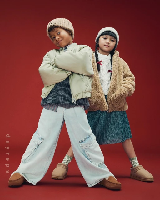 Two children wearing winter outfits pose against a red backdrop in a full shot. One child wears a beige jacket and blue jeans.