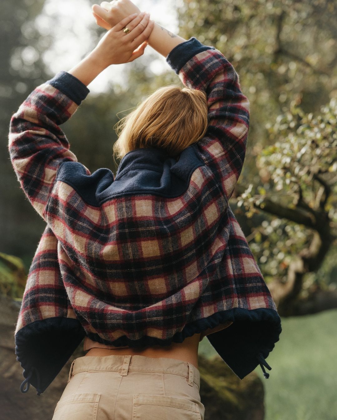 Woman in a plaid fleece jacket with arms raised, seen from behind in an outdoor park setting.