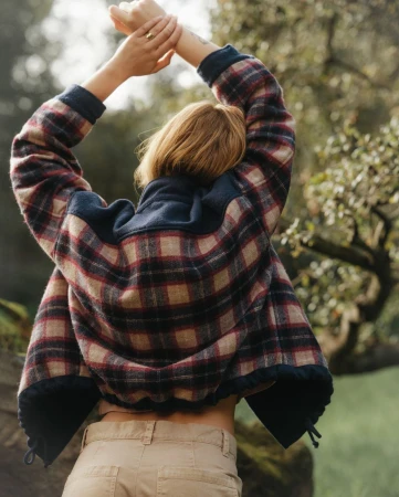 Woman in a plaid fleece jacket with arms raised, seen from behind in an outdoor park setting.