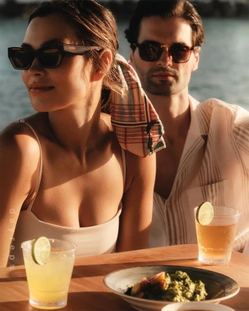 Man and woman wearing sunglasses sitting outdoors at a wooden table with cocktails and food, photographed by Wolfe + Von.