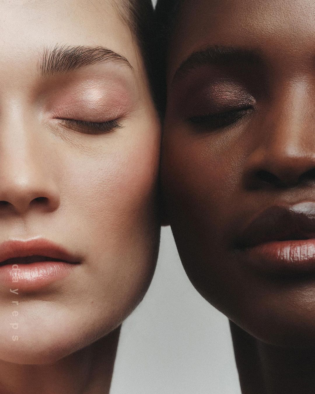 Close-up portrait of two female models with eyes closed pressing their faces together against a neutral background