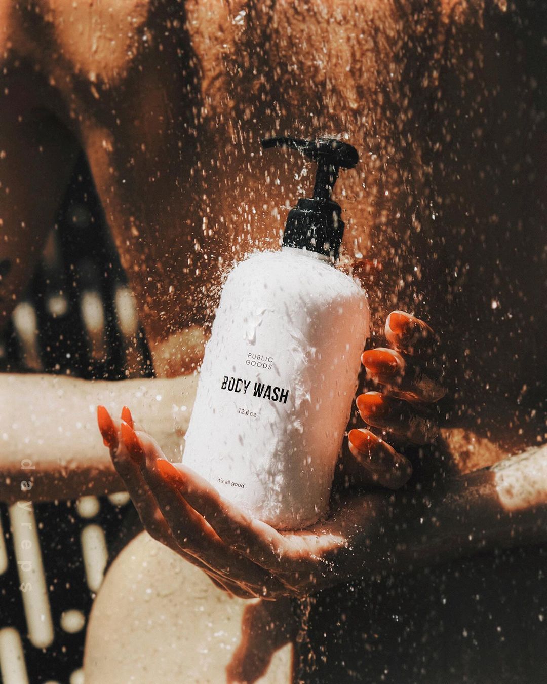 Close-up of a white bottle of body wash with a black pump dispenser held in a pair of hands as water droplets shower over it.