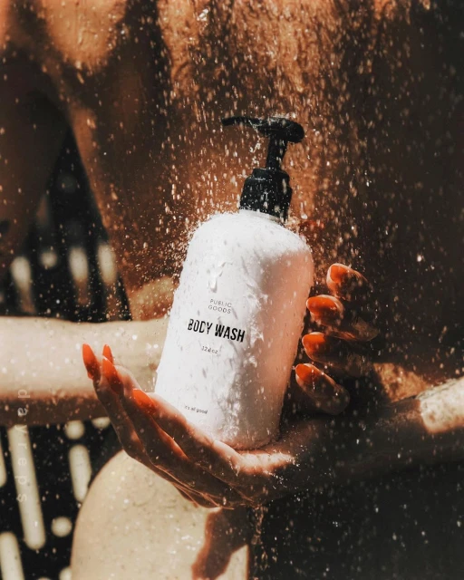Close-up of a white bottle of body wash with a black pump dispenser held in a pair of hands as water droplets shower over it.