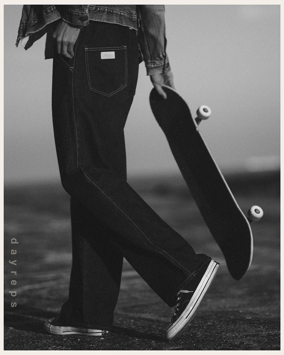 A person stands on a monochromatic urban surface wearing denim jeans and jacket, holding a skateboard, and wearing sneakers.