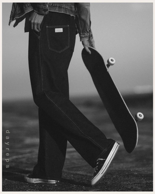 A person stands on a monochromatic urban surface wearing denim jeans and jacket, holding a skateboard, and wearing sneakers.