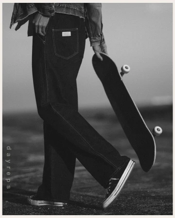 A person stands on a monochromatic urban surface wearing denim jeans and jacket, holding a skateboard, and wearing sneakers.