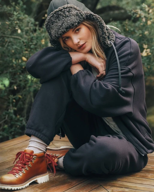 Woman wearing a navy sweat suit and fur-lined hat poses outdoors on a wood deck with green foliage behind her, fall fashion.