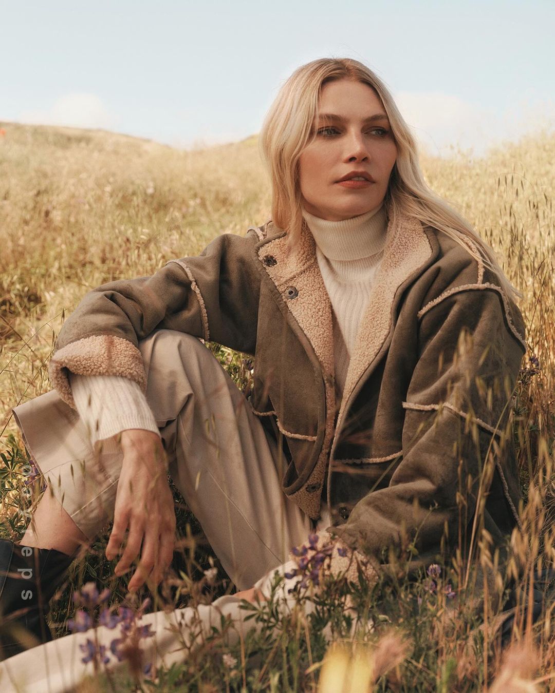 Blonde woman wearing an olive green shearling jacket and turtleneck sitting in a grassy meadow under warm sunlight.