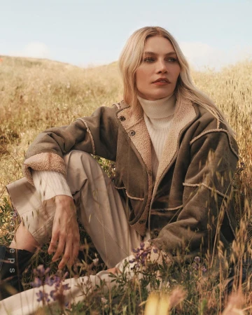 Blonde woman wearing an olive green shearling jacket and turtleneck sitting in a grassy meadow under warm sunlight.
