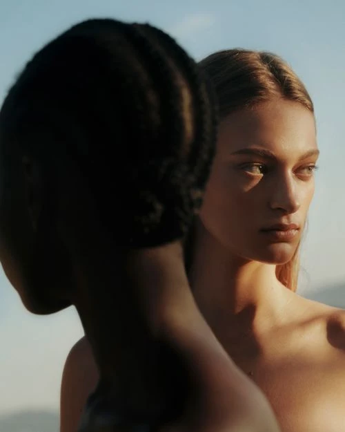Portrait of two women with different skin tones, one facing away, set against a light blue sky and distant landscape.