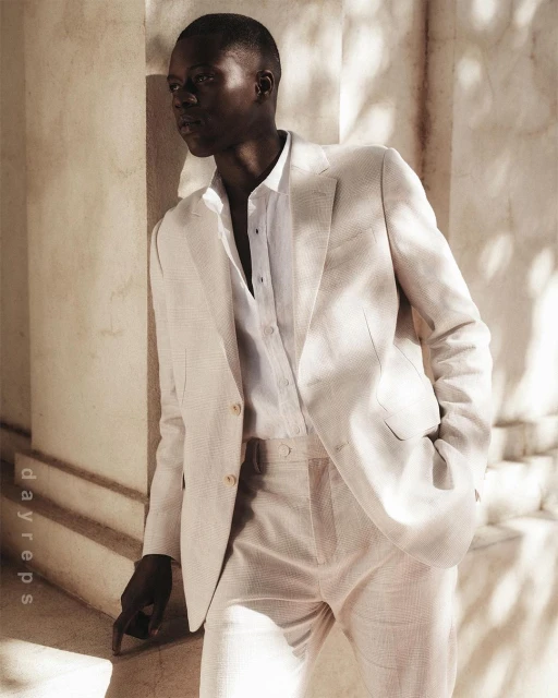 Fashionable man in a stylish linen suit poses indoors against a textured wall, showcasing a modern, sophisticated aesthetic in neutral tones.