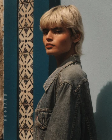 Portrait of a woman with blonde hair wearing a denim jacket standing against a blue wall with patterned tiles.