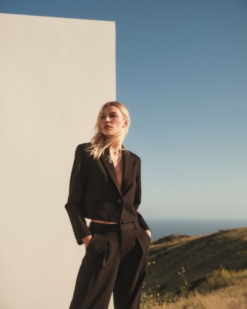Model wearing a black blazer and trousers posing outdoors against a white wall with a coastal landscape background.