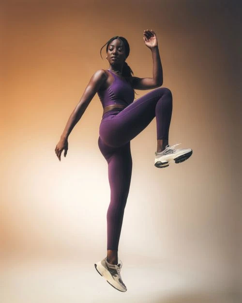 An athletic Black woman wearing a purple sports bra and leggings is mid-air with one leg tucked up, against an orange gradient background.