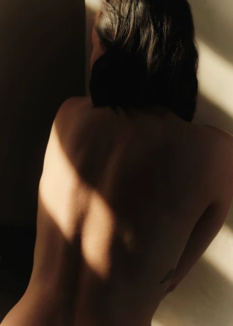 Back view of a partially undressed adult female bathed in warm sunlight against a plain wall. Focus on light and shadows.
