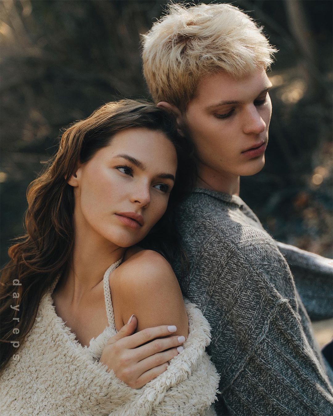 Two models, a blonde male and brunette female, pose closely together in a soft, natural outdoor setting, wearing knitted sweaters.