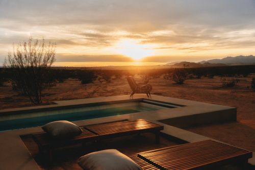 Sunset over desert landscape with lounge chairs near a pool at dusk, casting long shadows. Outdoor leisure and arid scenery combine.