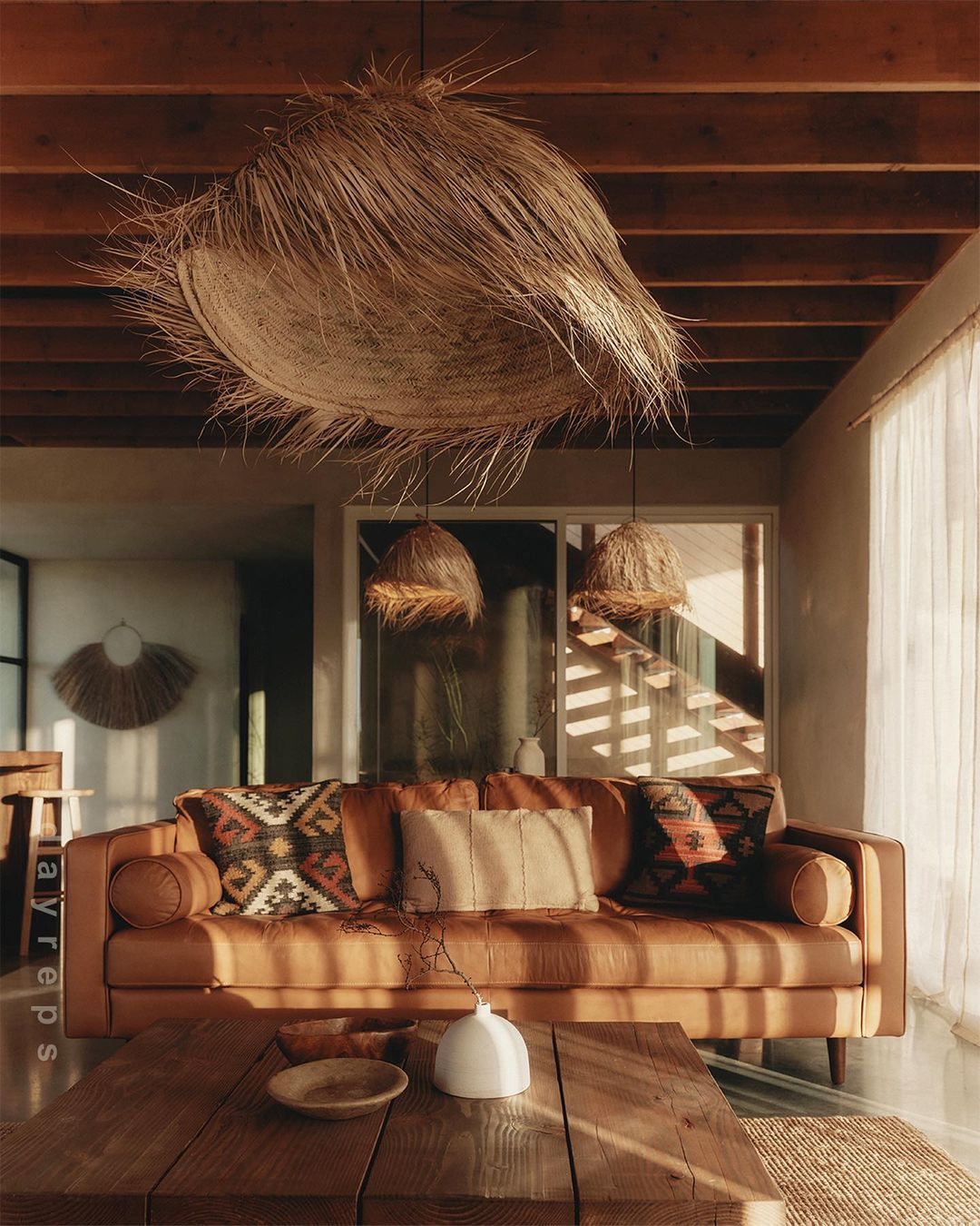 Living room interior features a brown leather sofa with patterned cushions, a wooden coffee table, straw light fixtures, and natural light.