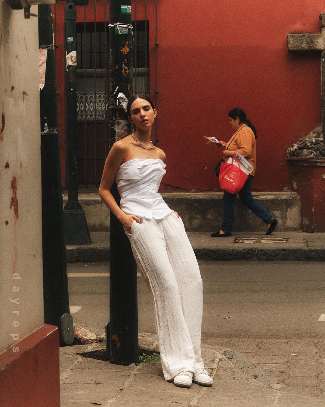 Young woman poses in a white top and pants near a city street corner with a red building. Woman in the background is out of focus.