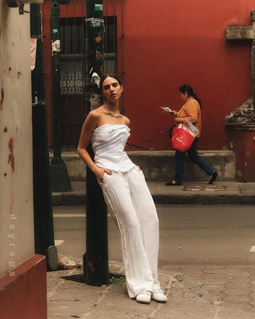 Young woman poses in a white top and pants near a city street corner with a red building. Woman in the background is out of focus.