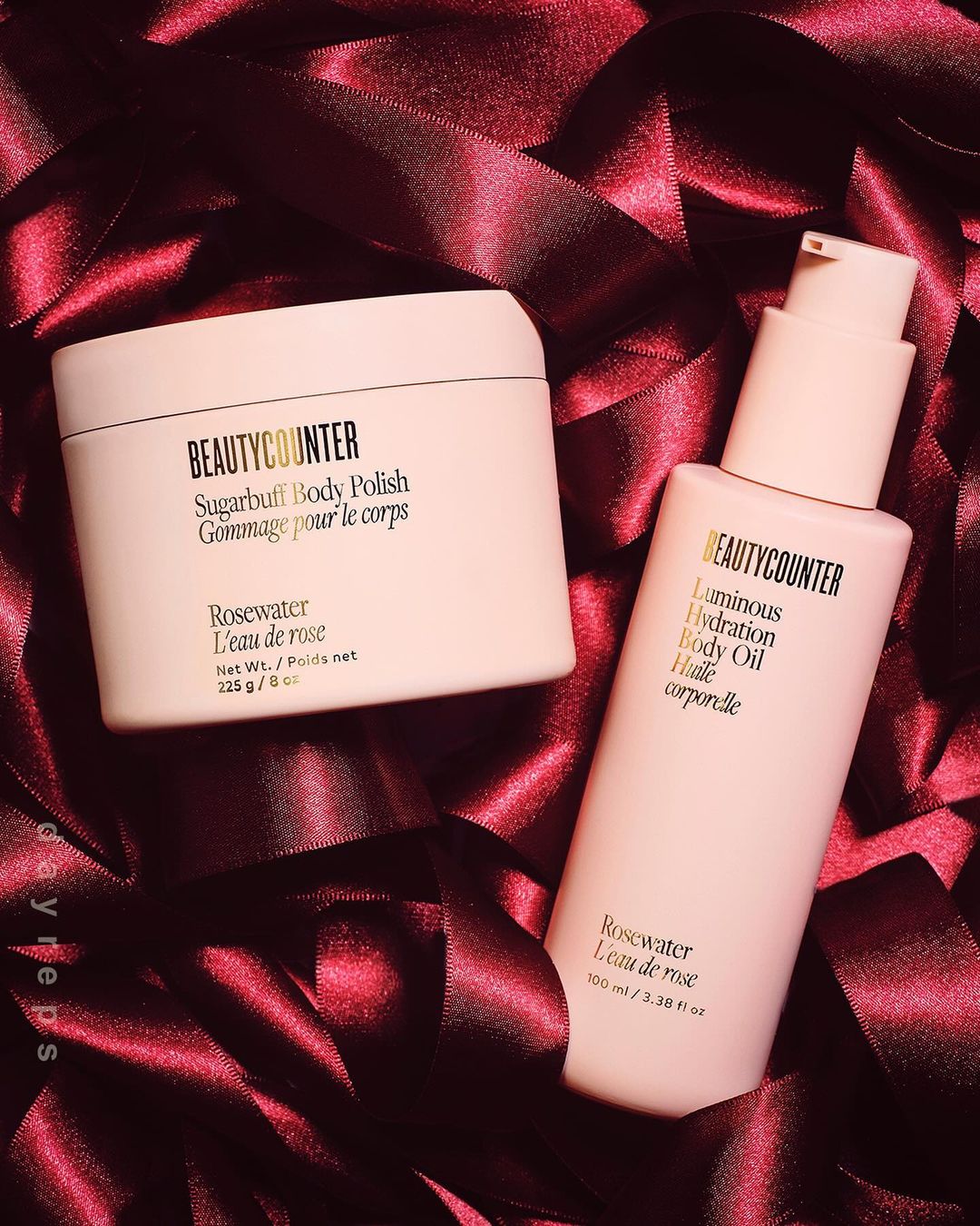 Two pink Beautycounter products, a body polish and body oil, are displayed on a background of dark pink satin ribbons, showcasing beauty and skincare.
