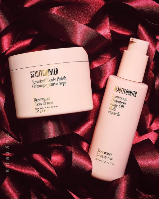 Two pink Beautycounter products, a body polish and body oil, are displayed on a background of dark pink satin ribbons, showcasing beauty and skincare.