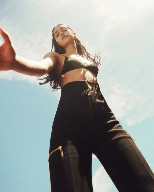Woman in black bra and high-waisted trousers poses with outstretched hand against a bright blue sky with a neutral facial expression.