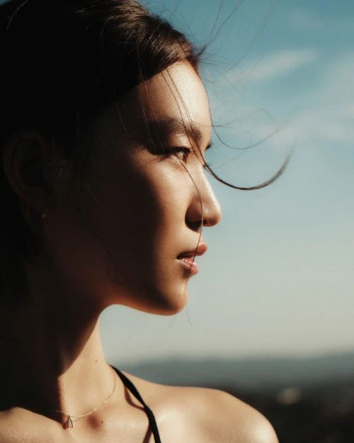 Side profile of a young Asian woman with windblown hair and a triangle pendant necklace, posing against a blurred blue sky and landscape.