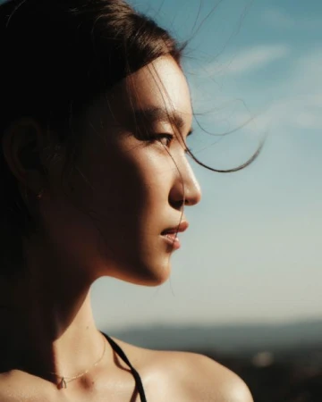 Side profile of a young Asian woman with windblown hair and a triangle pendant necklace, posing against a blurred blue sky and landscape.