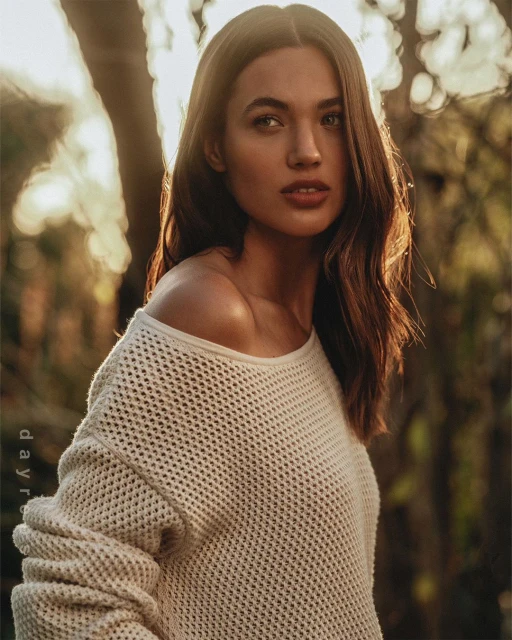 Attractive woman with long brown hair wearing a white sweater, posing outdoors in a forest setting with soft, warm light.