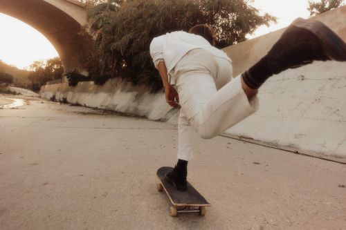 A person in white clothing is skateboarding in a cement riverbed under a bridge, with the late afternoon sun casting long shadows.