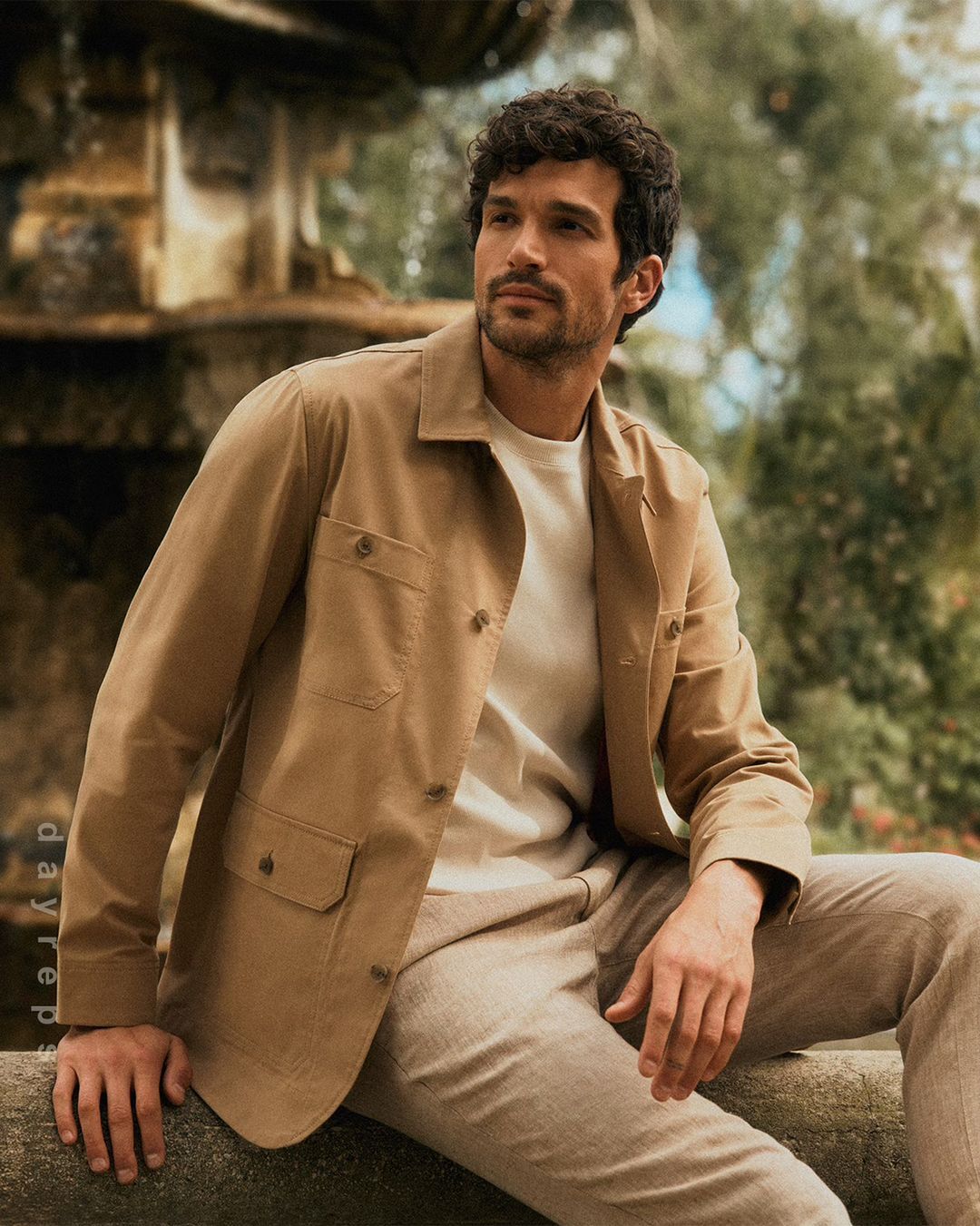 A man with dark hair and a tan jacket sits outdoors, looking off to the side with a natural expression, photographed in medium shot.