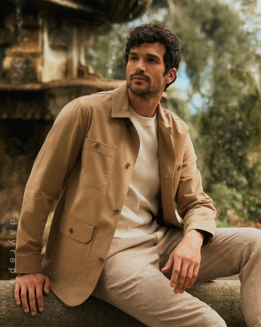 A man with dark hair and a tan jacket sits outdoors, looking off to the side with a natural expression, photographed in medium shot.