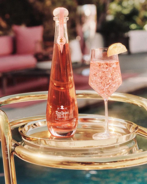 Rose-colored drink in glass with lemon slice and bottle of Don Julio Rosado Tequila on gold bar cart in an outdoor setting.