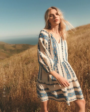 Blonde woman wearing a cream and blue patterned dress standing on a grassy hillside under a clear sky.