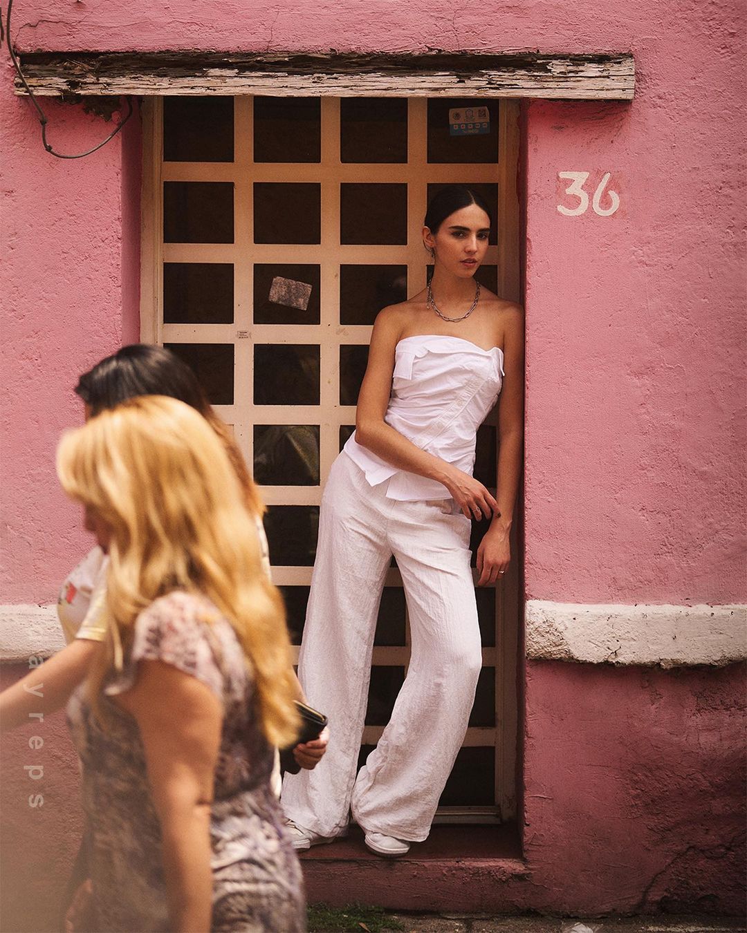 A woman in a white outfit poses by a pink building with a grid door while two women walk past in the foreground.