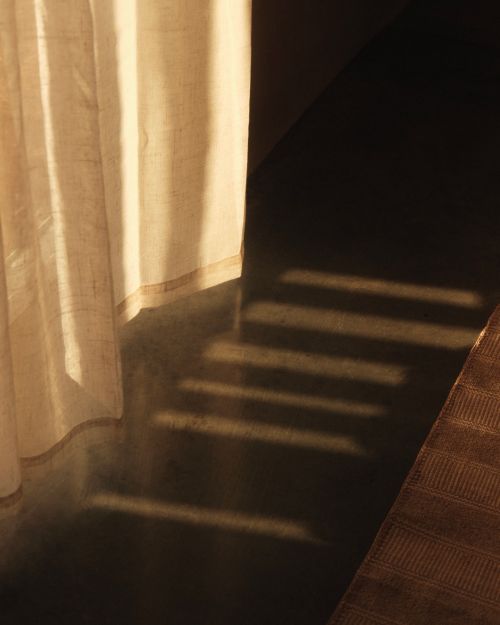 Sunlight streams into a room, casting light and shadow on a white curtain and dark floor, creating an abstract interior scene.