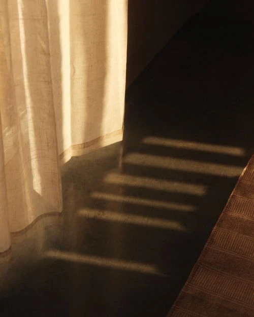 Sunlight streams into a room, casting light and shadow on a white curtain and dark floor, creating an abstract interior scene.