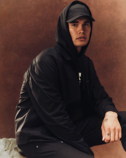 Young man in a black hoodie and baseball cap sits in a studio with a tan background, captured in a medium shot with natural lighting.