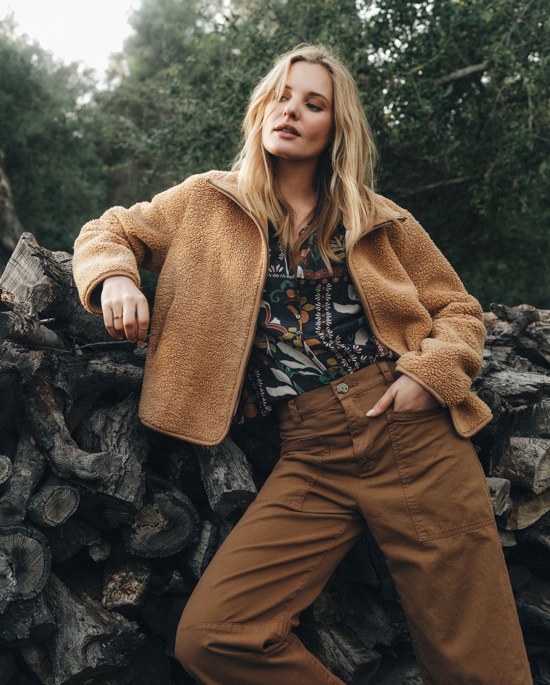 A young woman in casual wear, posing outdoors in a jacket and trousers, leans against a stack of wooden logs near a forest.