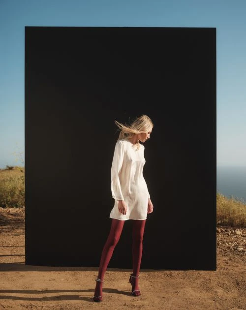 A slender blonde woman in a white dress and red tights stands on a hilltop near a black panel, with a distant ocean view and blue sky.