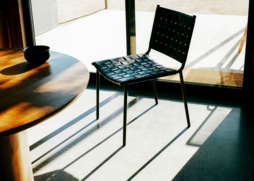 Black woven leather chair next to a round wooden table casting strong diagonal shadows on a concrete floor