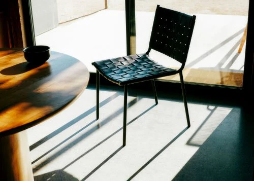 Black woven leather chair next to a round wooden table casting strong diagonal shadows on a concrete floor