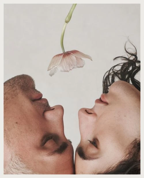 Artistic portrait of two people facing each other with closed eyes beneath a single flower with light pink petals on a white backdrop.