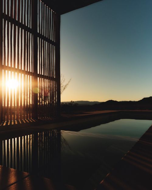 Sunset view through wooden slats over pool reflecting the sky, mountains in the distance, outdoor dusk scene.