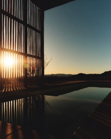 Sunset view through wooden slats over pool reflecting the sky, mountains in the distance, outdoor dusk scene.