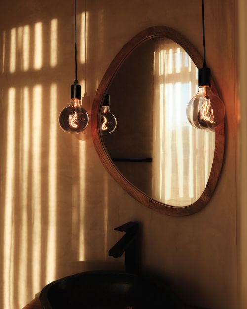 Illuminated light bulbs hang over a black sink and faucet, reflected in a mirror with warm lighting and window shadows.