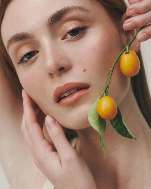 Close-up portrait of a woman holding a small leafy branch with two kumquat fruits near her face.
